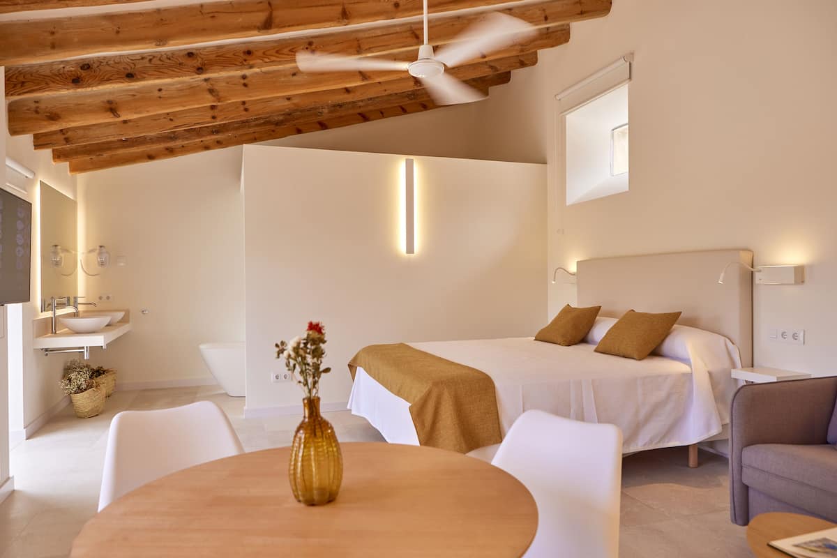 A modern hotel room with a bed, a round table with a vase, a sofa, a ceiling fan, and a bathroom with a sink and a bathtub.