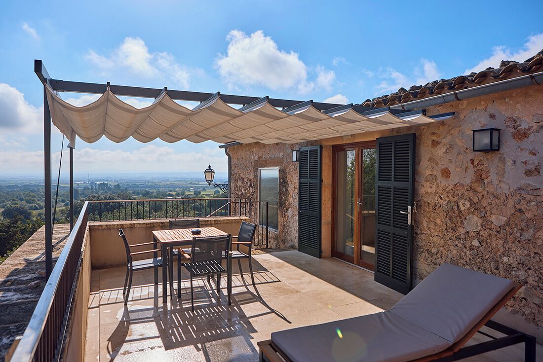 Una terraza con mesa y sillas, una silla de descanso, una pérgola y un muro de piedra con contraventanas.