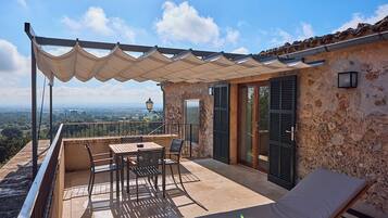 Una terraza con mesa y sillas, una silla de descanso, una pérgola y un muro de piedra con contraventanas.