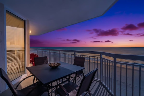 Un balcon avec une table et des chaises, une vue sur l’océan et un ciel au coucher du soleil.