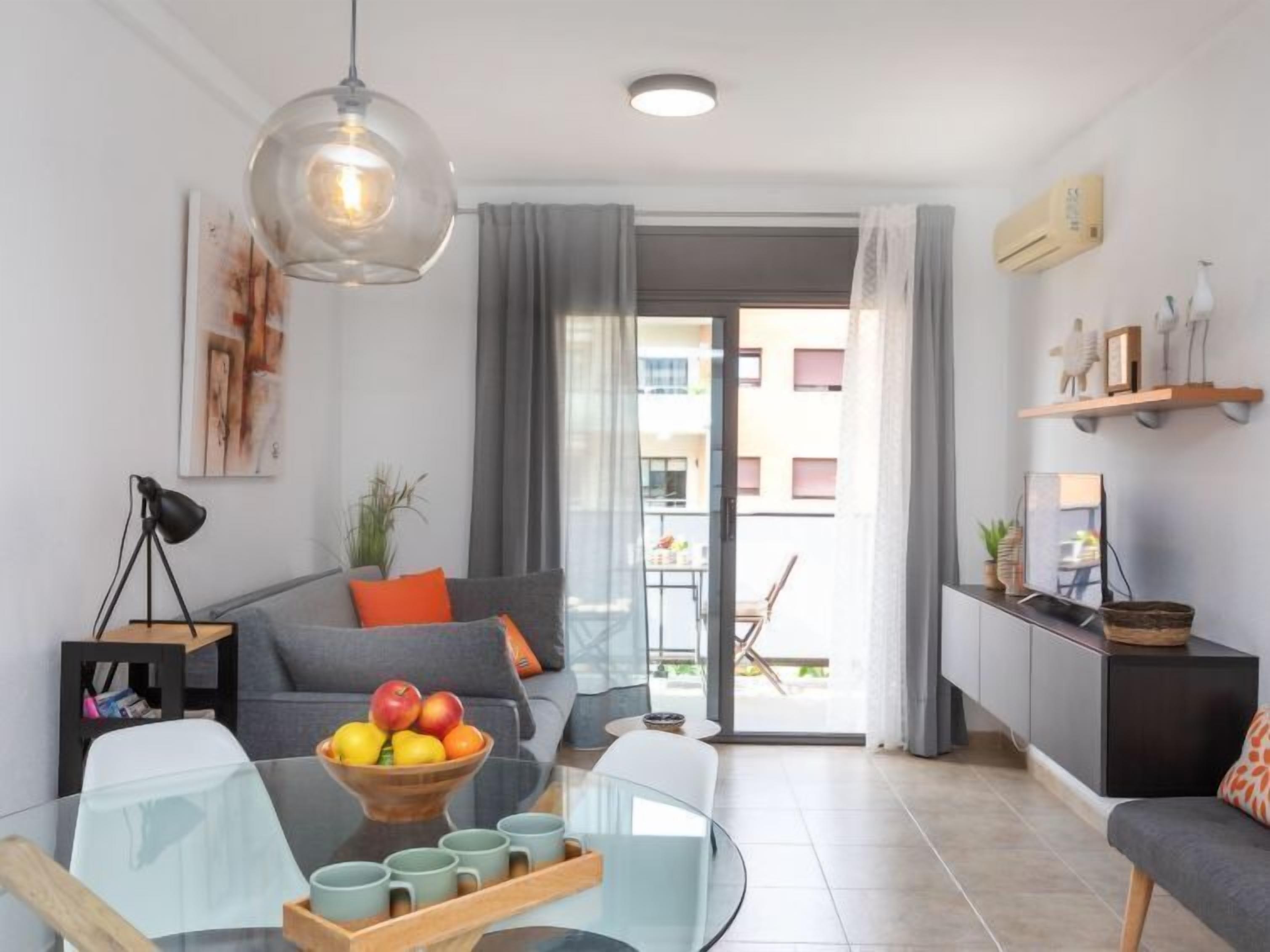 Ein modernes Wohnzimmer mit grauem Sofa, einem Glastisch und einer Obstschale. Es gibt einen Essbereich mit weißem Tisch und Stühlen sowie eine Küchenzeile mit einem schwarzen Schrank und einer Hängeleuchte.