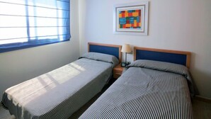 Une chambre avec deux lits simples, une fenêtre avec des stores, une table de chevet avec une lampe et une peinture abstraite aux couleurs vives accrochée au mur.