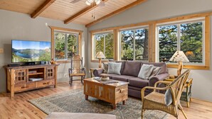 A cozy living room with a wooden ceiling, large windows with a view of trees, a television, a wooden cabinet, a brown sofa, and wicker chairs.