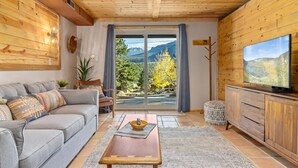 A cozy living room with a grey sofa, wooden coffee table, and a TV on a wooden cabinet. A large window offers a view of mountains and trees.