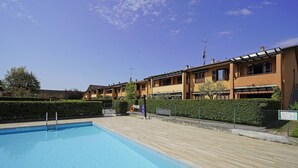 A swimming pool with a wooden deck, surrounded by apartment buildings with balconies and a clear blue sky.