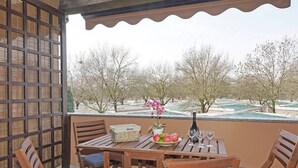 A wooden table with chairs and a bowl of fruit, a bottle, and glasses on a balcony with a view of trees and a pool.