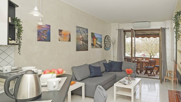 A modern living room with a grey sofa, white coffee table, and a dining area with a kettle and plates.