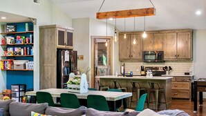 A modern kitchen with wooden cabinets, a central island, and bar stools. There is a dining area with a table and chairs, and a living room with a sofa.