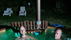 A hot tub with two people in it, surrounded by a wooden deck and lounge chairs. - House of Rei With Hot Tube (Ocolis)