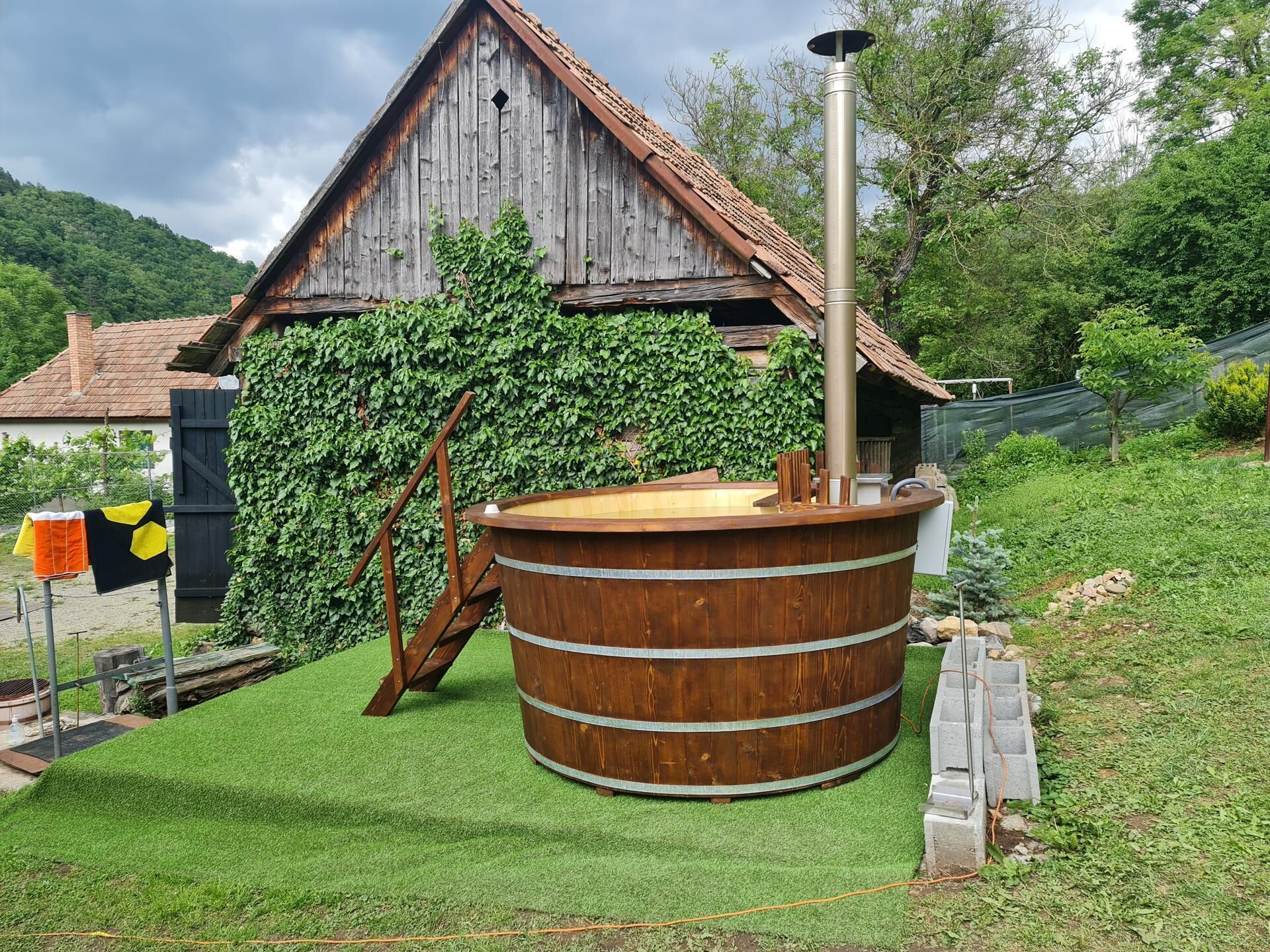 A rustic wooden sauna with a metal chimney, surrounded by a well-maintained lawn and a wooden structure with a tiled roof.