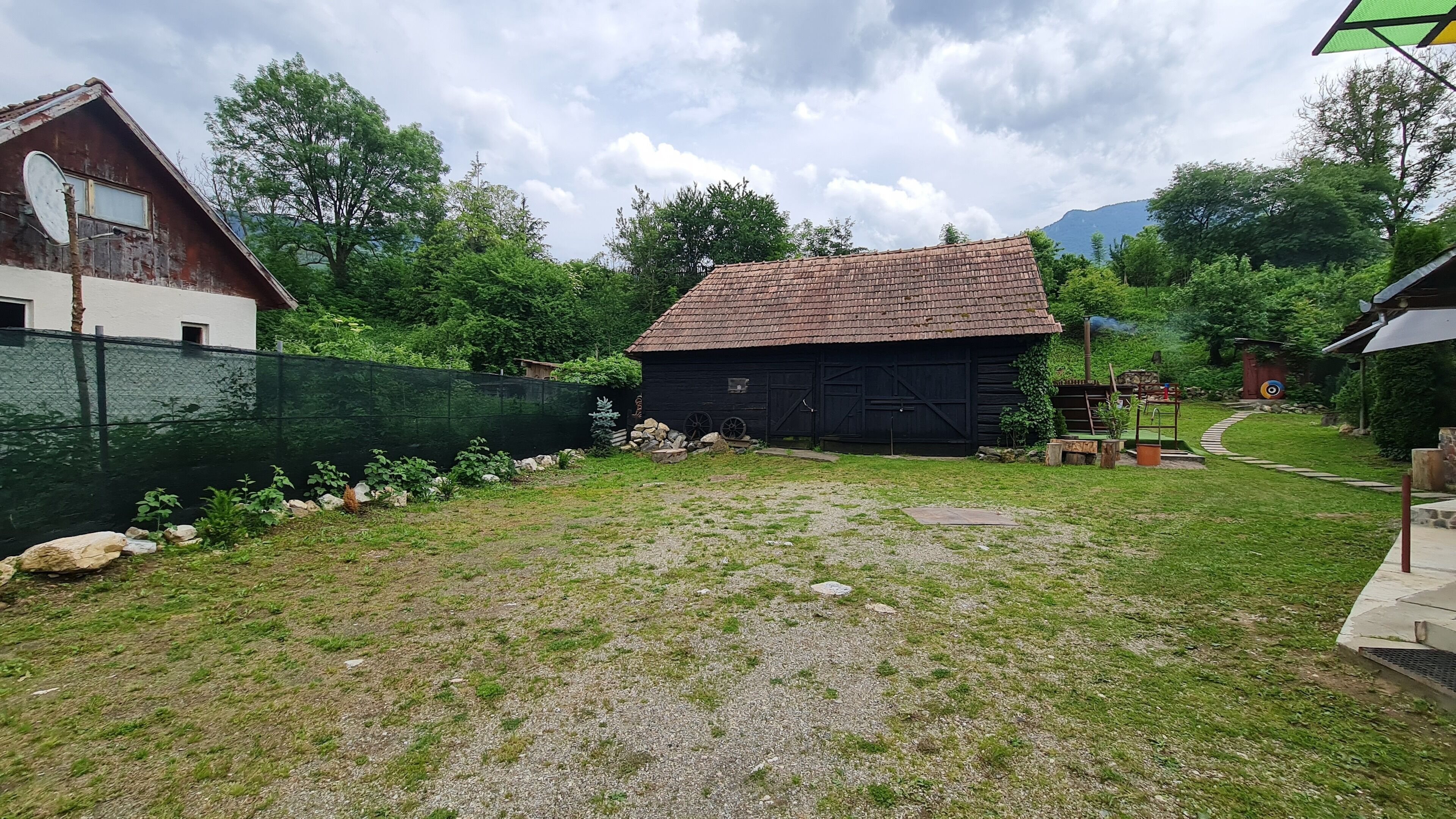 A rustic wooden cabin with a thatched roof, surrounded by a gravel yard and lush greenery.