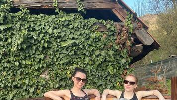 Two people relaxing in a wooden hot tub with a rustic wooden building and ivy-covered walls in the background.
