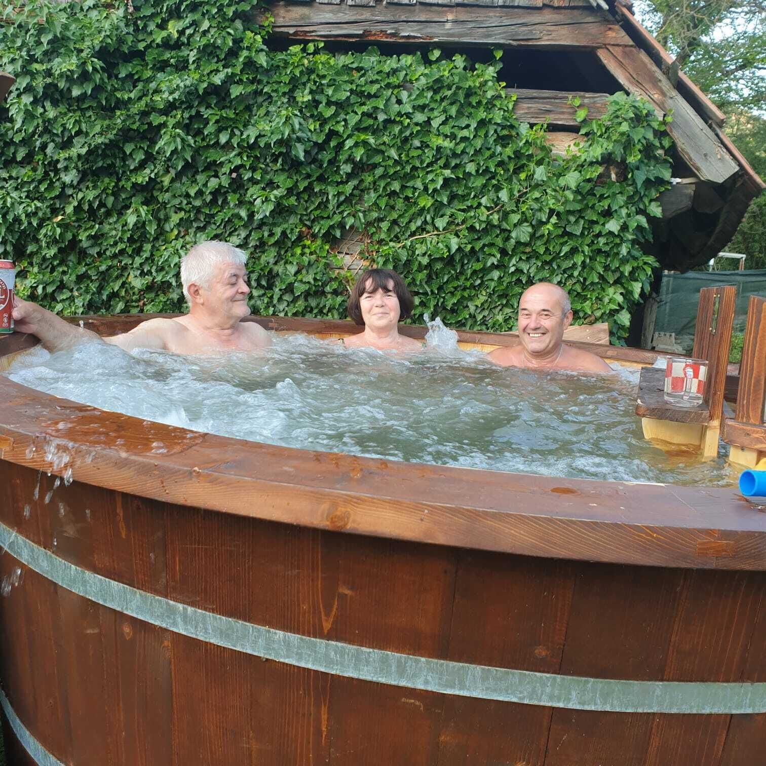 A wooden hot tub with three people in it, surrounded by a wooden structure and a green ivy-covered wall.