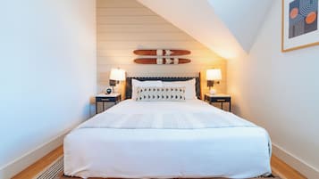 Une chambre à coucher bien aménagée, avec un lit placé au centre, sous un plafond en pente, flanqué de deux tables de chevet avec des lampes, d’une planche de skate fixée au mur et d’une œuvre d’art abstraite encadrée.