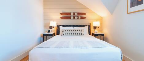 A neatly arranged bedroom with a bed centered under a sloped ceiling, flanked by two bedside tables with lamps, a wall-mounted skateboard, and a framed abstract artwork.