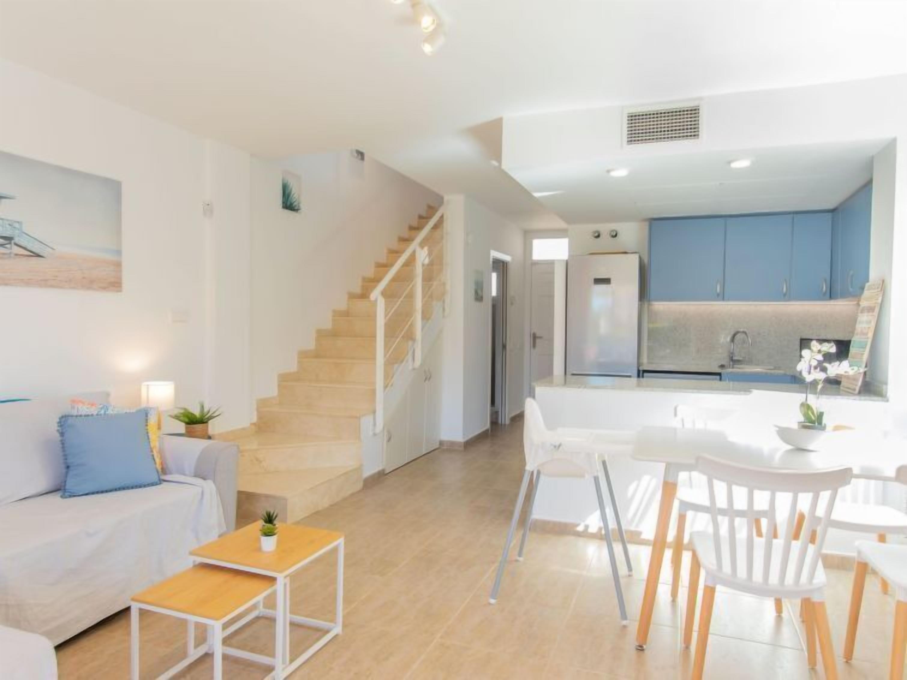 A modern living room with a staircase, a dining area with white chairs and a table, and a kitchen with blue cabinets.