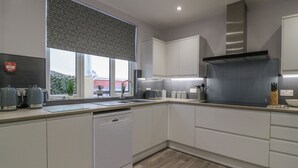 Eine moderne Küche mit weißen Schränken, einem Geschirrspüler, einem großen Fenster mit gemustertem Rollo und einer grauen Dunstabzugshaube.