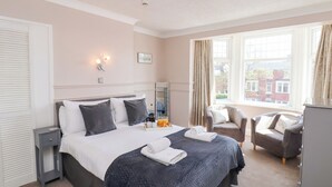 Ein Hotelzimmer mit einem großen Bett, einem Nachttisch, einem Sessel, einem Schreibtisch und einem Fenster mit Vorhängen.