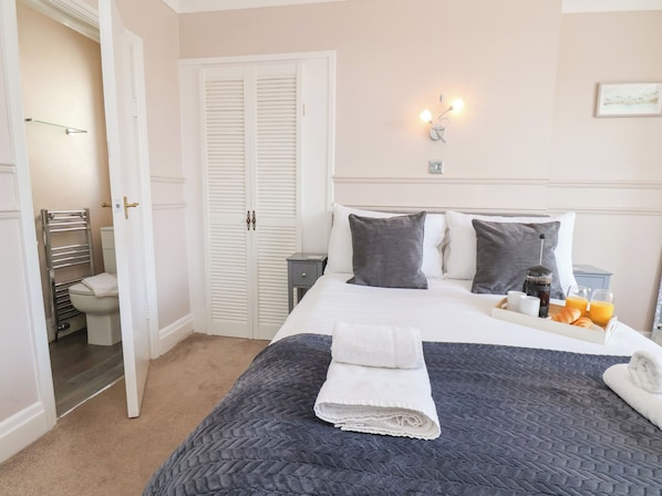 A neatly arranged bedroom with a bed, bedside tables, a nightstand, a wardrobe, and a bathroom visible through an open door.