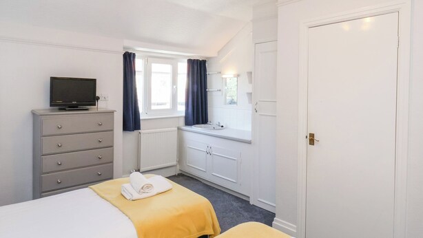 A hotel room with a bed, a TV on a dresser, a window with curtains, a bathroom with a sink, and a door. - North Beach Lodge (Scarborough)