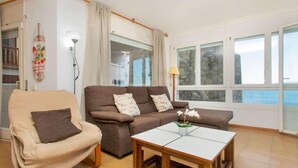 Ein Wohnzimmer mit einem beigen Sessel, einem braunen Sofa und einem Couchtisch aus Holz. Vor großen Fenstern bietet sich Blick auf das Meer, ergänzt durch Vorhänge und eine Lampe.