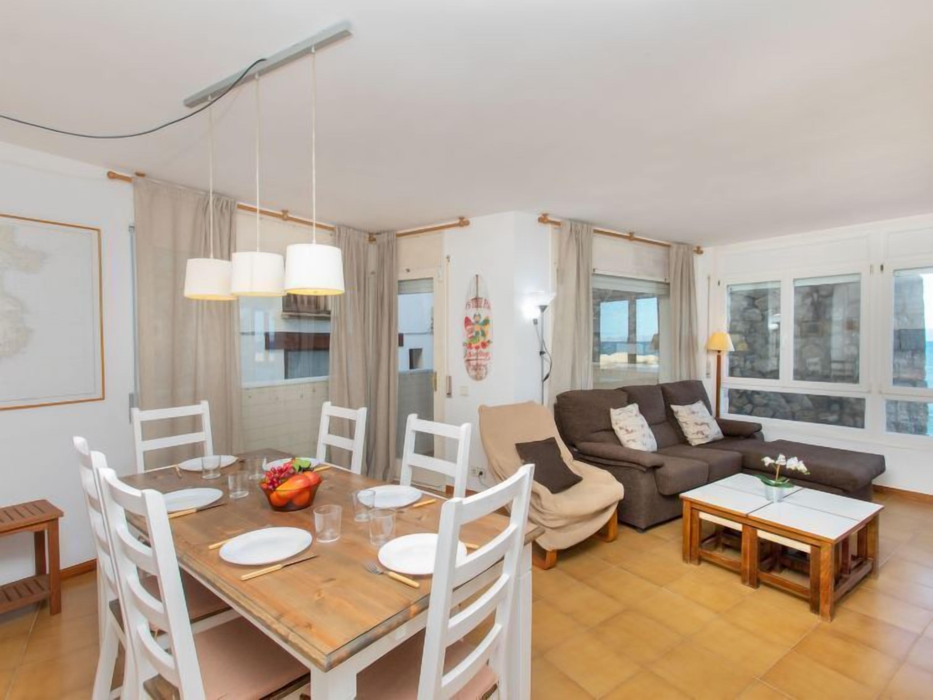 A spacious living area with a wooden dining table set for four, surrounded by white chairs. A brown sofa and a small wooden table with a lamp are in the living room.