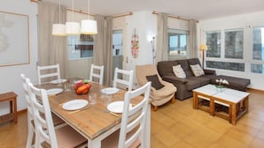 Ein geräumiger Wohnbereich mit einem Holzesstisch, gedeckt für vier Personen, umgeben von weißen Stühlen. Ein braunes Sofa und ein kleiner Holztisch mit einer Lampe befinden sich im Wohnzimmer.