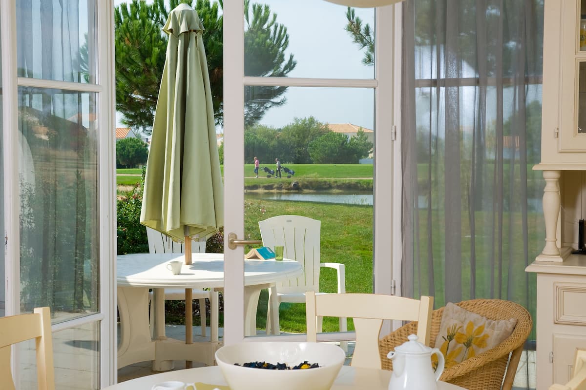 Une pièce comprenant une table à manger, des chaises, un fauteuil en osier, un panier de fruits et une vue sur un espace verdoyant avec des arbres et un étang.