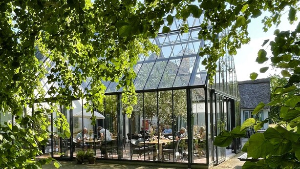 Et glashuset udendørs spiseområde med borde og stole, omgivet af grøn vegetation og en moderne bygning i baggrunden.