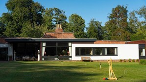 A modern house with a large lawn, a tennis court, and a patio area with outdoor furniture.