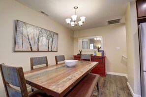 A dining area with a wooden table, chairs, a large painting, a chandelier, and a refrigerator.