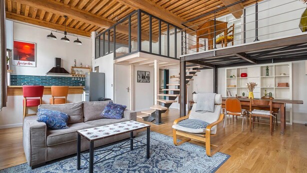 Amplia sala de estar con techo alto, vigas de madera, una escalera y una zona de comedor con mesa y sillas.