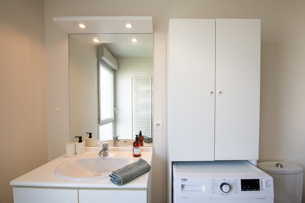 Un baño moderno con lavamanos, espejo y mueble blancos, lavadora integrada y un inodoro.