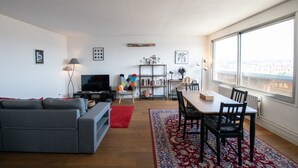 A modern living room with a grey sofa, a TV, a colorful chair, a dining table with chairs, and a large window with a city view.