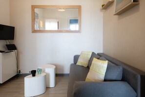 Een moderne woonkamer met een grijze bank, twee witte bijzettafels, een flatscreen televisie en een spiegel aan de muur.