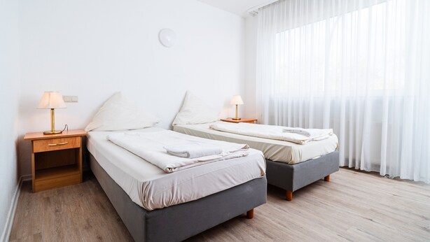 A hotel room with two single beds, wooden nightstands with lamps, a small wooden shelf, and a large window with sheer curtains. - G & L Hotel (Dortmund)