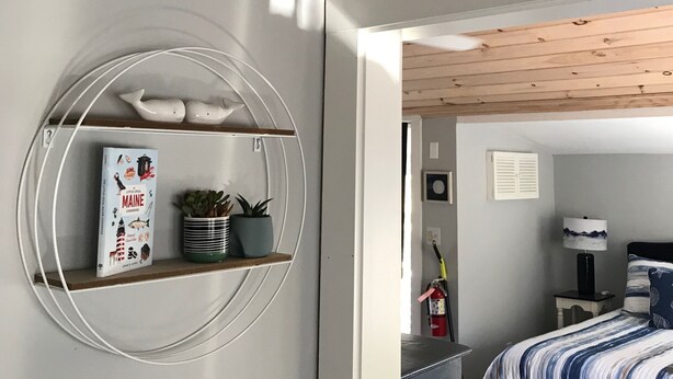 Une pièce avec une étagère circulaire sur laquelle sont posés un livre et des plantes, un panneau indiquant « PLAGE » sur le mur, et un lit avec une literie à rayures.