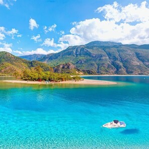 A clear view of a lake with a boat, surrounded by mountains and a small beach area with structures. - Ideally located 4 bedroom villa with private pool (Muğla)