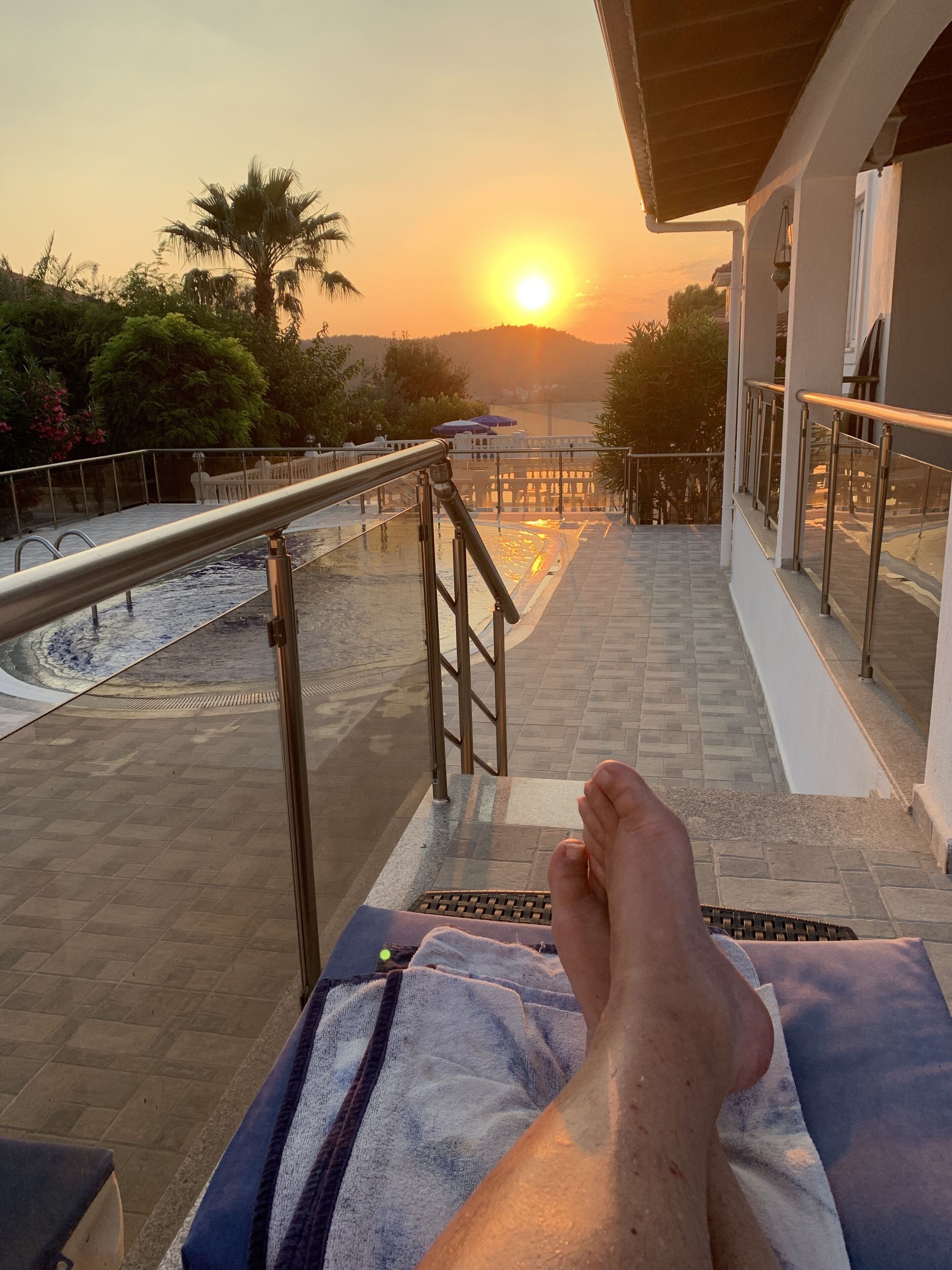 A balcony with a view of the sunset, a pool, and a staircase leading down.