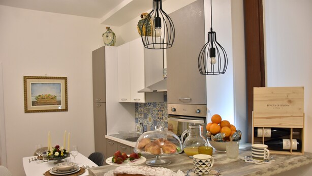 Una cocina moderna con armarios blancos, una mesa de comedor dispuesta con platos, vasos y un pastel. Hay luces colgantes, un frutero y una estantería para vinos.