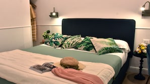 Una cama impecable con colcha verde y blanca, una manta rosa y un sombrero de paja. Hay dos almohadas con estampado de hojas tropicales, una peque ña planta en maceta y una lámpara de pared.