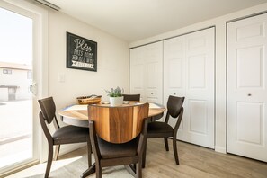 Un coin repas avec une table ronde en bois, quatre chaises, une petite plante en pot et un panier. Un panneau est accroché au mur, et on aperçoit un ensemble de portes blanches à l’arrière-plan.