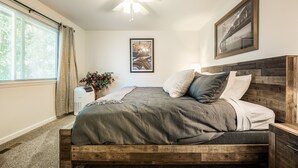 Un dormitorio con estructura de cama de madera, ventilador de techo, ventana con cortinas, purificador de aire montado en la pared y un cuadro enmarcado.
