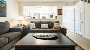 Una sala de estar con sofá, mesa de centro y lámpara. Se observa una cocina al fondo con armarios y microondas.