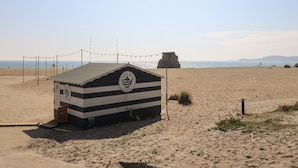 Eine kleine, gestreifte Strandhütte mit einem Logo, auf einem Sandstrand mit freiem Blick auf das Meer und die entfernten Hügel.