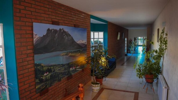 A hallway with a large painting of a mountainous landscape, potted plants, and a view into another room. - Hotel Ariel (El Calafate)