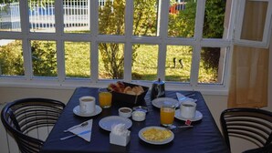 A breakfast table set with plates, cups, and a basket of bread, viewed through a window with a view of a garden. - Hotel Ariel (El Calafate)