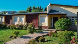 A two-story building with a yellow facade and a red brick section. There is a small garden with various plants and flowers in front of the building. - Hotel Ariel (El Calafate)