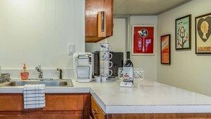 Cozinha com bancada branca, pia de aço inoxidável e armários de madeira. Há diversos eletrodomésticos, incluindo uma cafeteira e uma torradeira. A parede exibe várias obras de arte emolduradas e um cartaz vermelho.