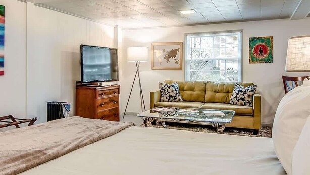 Quarto de hotel com cama, sofá, mesa de centro, televisão, cômoda e janela com persianas.